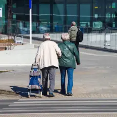 Starsza para idzie przez pasy, mężczyzna ciągnie wózek. To obraz codzienności, gdzie każdy krok to dowód na to, że życie trwa, a stopa zastąpienia nie jest jeszcze potrzebna.