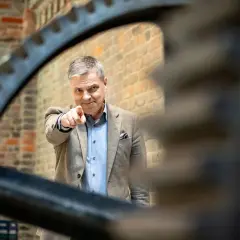 Mężczyzna w garniturze wskazuje palcem, jakby prezentował zestawienie finansowe. Za nim ceglana ściana i zębate koło.