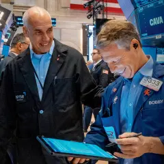 Dwóch mężczyzn w kurtkach NYSE śmieje się, patrząc na tablet. Jeden z nich, Bobby, pokazuje coś na ekranie, a drugi, z napisem "skup akcji własnych" na piersi, uśmiecha się szeroko.