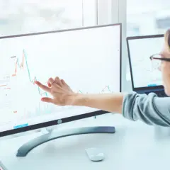 Kobieta analizuje wykres Sunex na monitorze, wskazując palcem na trend wzrostowy. Obok leży tablet z podobnymi danymi.