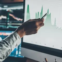 Dłoń z długopisem wskazuje na wykres świecowy, analizując siłę relatywną trendów na ekranie monitora.