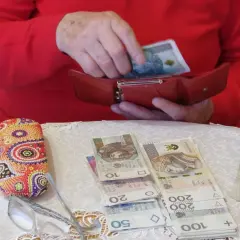 Dłonie chowają banknot 100 zł do czerwonego portfela. Obok leżą pieniądze, okulary i etui. To pokazuje, gdzie trzymać pieniądze.
