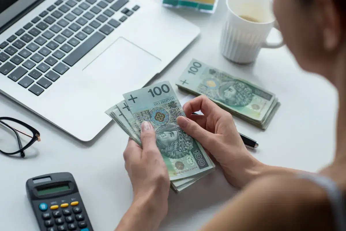 Ręce liczą banknoty 100 zł, obok laptop, kalkulator i okulary. Czas pomyśleć, jak pomnożyć 100 zł.